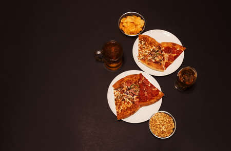 Slices of pepperoni pizza, ham pizza and chicken pizza on white plates, glass of beer, mug of beer, peanuts and potato chips on dark background. Top view, copy space.の写真素材