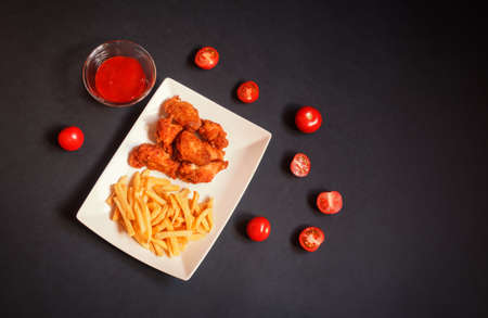 Chicken wings with french fries and fresh ripe tomatoes on dark background. Top view, copy space.の写真素材
