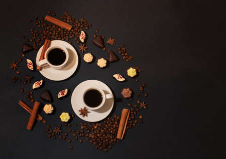 Two white cups of coffee, coffee beans, spices and chocolate candies on dark background. Top view, copy space.の写真素材