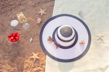 Hat, sunglasses, glass of champagne, fresh strawberry, starfishes and seashells on towel on sand...