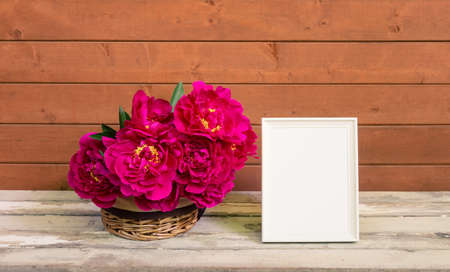 Red peony flowers in wicker basket and empty white frame on wooden table. view with copy space.の写真素材