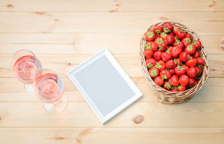 Two glasses of rose wine, white frame and ripe red strawberries in wicker basket on wooden table. Top view, copy space.の写真素材