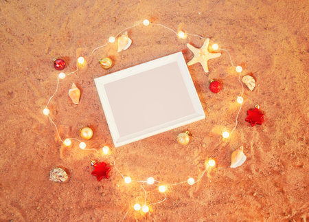 White frame, chrismas decorations, glowing christmas lights, starfishes and seashells on sand beach. Top view with copy space. Beach christmas and New Year celebration concept.の写真素材