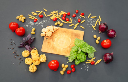 Cutting board, fresh tomatoes and onions, raw penne and fettuccine, cheese, basil, green olives and spices on dark background. Top view, copy space.の写真素材