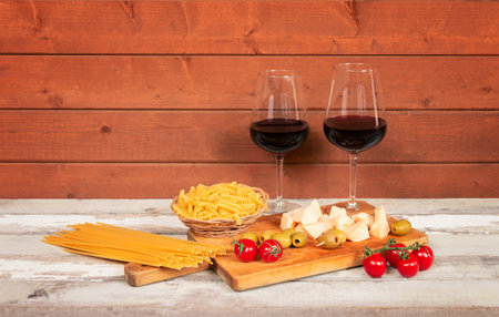 Two glasses of red wine, raw pasta fusilli and spaghetti, cherry tomatoes, green olives and pieces of cheese on aged wooden table.の写真素材