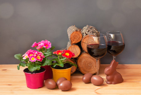 Easter concept. Primula flowers, glasses of red wine, chocolate easter eggs, chocolate bunny and pile of firewood on wooden table. view with copy space.の写真素材