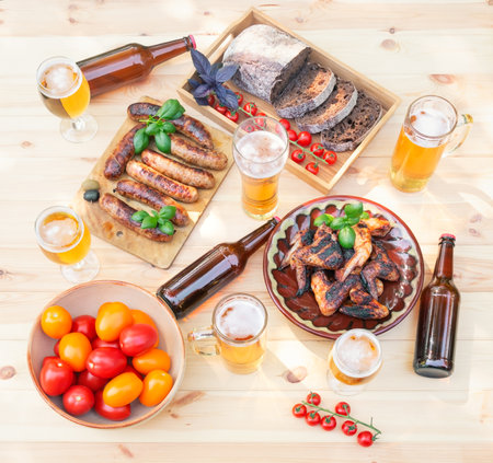 Grilled chicken wings, grilled sausages, mugs and glasses of beer, beer bottles, rye bread and clay bowl with red and yellow tomatoes on wooden table. top view.の写真素材