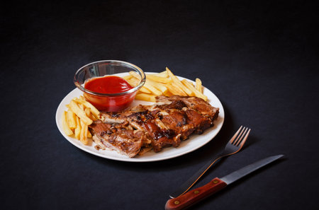 Baked pork ribs with french fries and tomato ketchup on white plate on dark background. selective focus.の写真素材