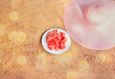 Large pink summer hat and plate with pieces of watermelon on sand beach. Beach summer holidays concept. Top view, copy space.の写真素材