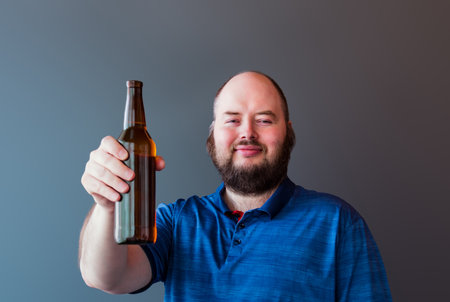 Cheers! Portrait of a young man wearing a blue t-shirt holding a bottle of beer. gray background. Selective focus on face.の写真素材