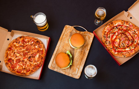 Burgers on cutting board, pizzas in boxes, mug and glasses of beer on dark background. top view.の写真素材