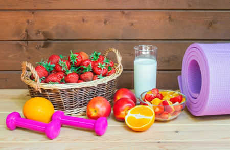 Fresh strawberry, yogurt, healthy fruit salad made of strawberry, oranges and nectarines, fresh oranges and nectarines near, dumbbells and yoga mat on wooden surface.の写真素材