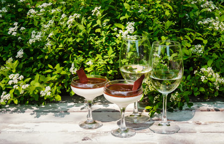 Homemade delicious italian dessert panna cotta with chocolate syrup and two glasses of white wine on rustic wooden table on fresh white Spiraea flowers backgroundの写真素材