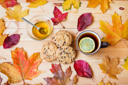 Cup of tea with lemon, honey in glass bowl, cookies with chocolate drops, and colorful autumn leaves on wooden table. top view.の写真素材