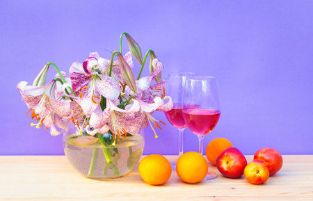 Bouquet of white lilies with purple spots in glass round vase, glasses of rose wine and fresh fruits on wooden table. purple background. view with copy space.の写真素材