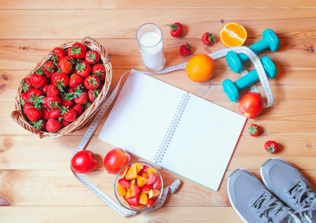 Empty notebook, yogurt, healthy fruit salad made of strawberry, oranges and nectarines, fresh oranges and nectarines near, wicker basket of fresh strawberry, dumbbells, sneakers and measuring tape on wooden surface. Top view with copy space.の写真素材