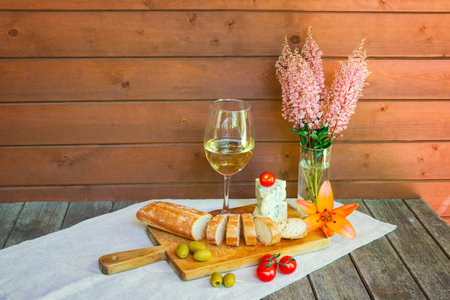 Glass of white wine, blue cheese, baguette, cherry tomatoes and olives, lily and astilbe flowers on rustic wooden table.の写真素材