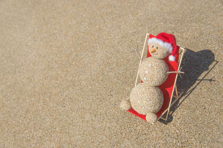 Sandy snowman in red santa hat sunbathing in beach lounge. Holiday concept for New Years and Christmas Cards.の写真素材