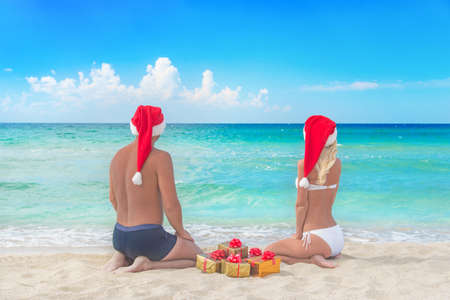 Lovers couple in red santa hats and many golden packed presents on sea beach - christmas vacation conceptの写真素材
