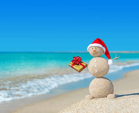 Smiley sandy snowman at beach in christmas hat with golden gift. Holiday concept for New Years Cards.の写真素材