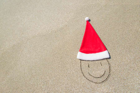 Santa Claus hat and smiley face on seashore against waves and blue sky - winter vacation conceptの写真素材