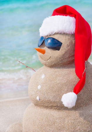 Sandy christmas snowman in red santa hat and sunglasses at sunny beach. Holiday concept for New Years Cards.の写真素材