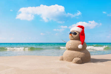 Sandy christmas snowman in red santa hat and sunglasses at sunny beach. Holiday concept for New Years Cards.の写真素材