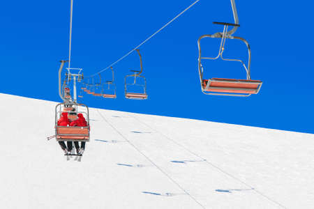 Skiers couple in Christmas Santa hats and snowboarders at ski lift transport up a snowy downhill at a winter ski resort in the Alps, white snow and blue sunny sky backgroundの写真素材