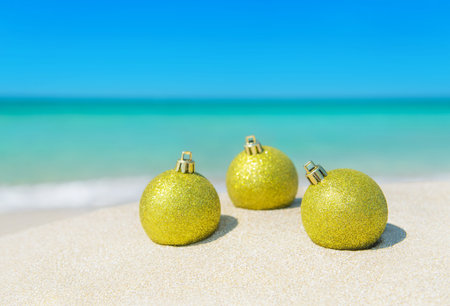 Three golden christmas tree balls decorations shining at sun on tropical ocean beach sand  - winter...