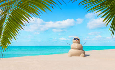 Positive sandy snowman in black sunglasses and carrot nose at sunny tropical ocean palm beach. Happy New Year and Merry Christmas travel destinations for tropical vacations conceptの写真素材