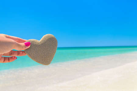Heart made of sand in womans hand against tropical ocean beach background. Romantic, love, honeymoon, wedding or Saint Valentines day greeting cards concept.の写真素材
