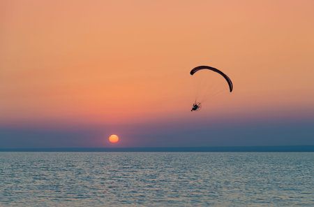 Silhouette of powered paraglider soaring flight over the sea against marvellous orange sunset sky....