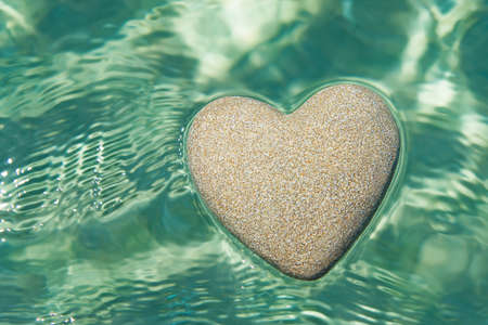 Heart made of sand floating in transparent turquoise ocean ripples water background, Saint Valentines day greeting cards, romantic love, honeymoon, proposal or wedding concept.の写真素材