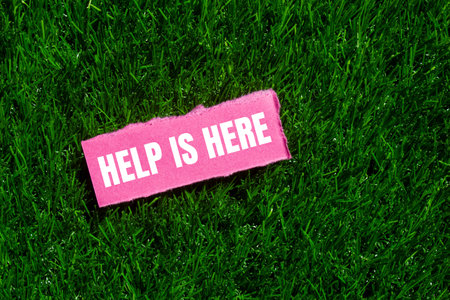 Help is here message written on ripped torn pink paper piece on green grass background. Conceptual help is here symbol. Copy space.の写真素材