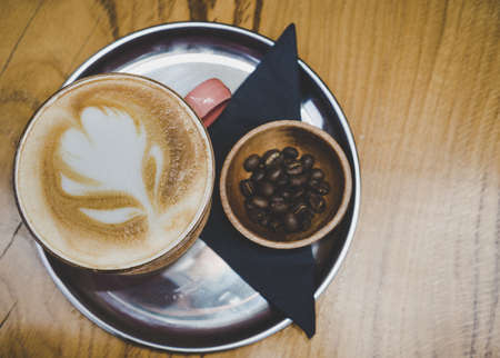 Fresh cappuccino on metal plate on wooden surface. Decorated with coffee beans in small wooden bowl. With flower art on foam.の写真素材