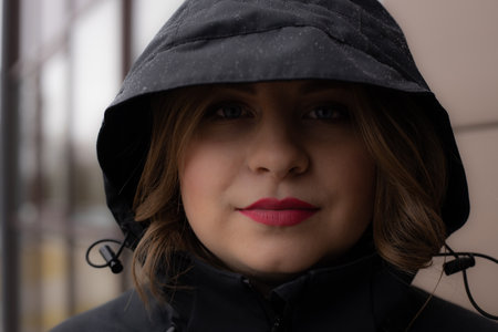 A woman with red lips in a black hoodie under the rain. Beautiful young woman under the hood. Young girl with beautiful red lipsの写真素材