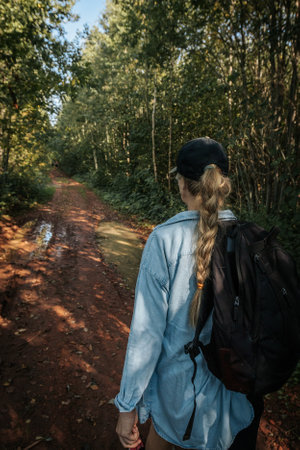 A young woman with a backpack walks in the forest on a dirt road.
