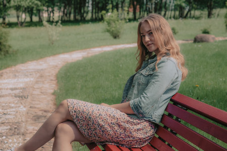 Beautiful girl sitting on a bench in the park. The girl is dressed in a denim jacket.の写真素材