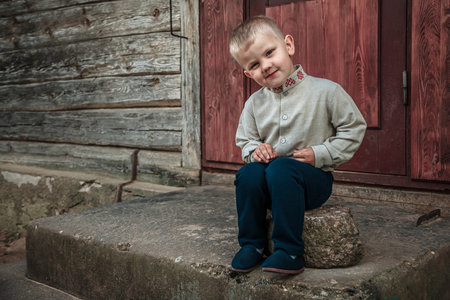 Portrait of a little boy sitting on a stone in front of a wooden houseの写真素材