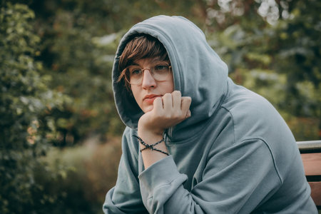 A young woman in a gray hoodie and glasses sits on a bench in the park.の写真素材