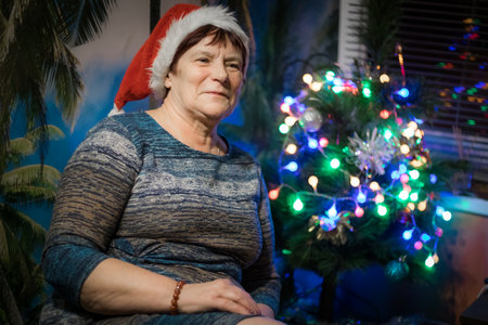 Mature woman in santa hat sitting near christmas tree at homeの写真素材