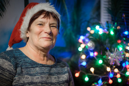 Portrait of an elderly woman in a Santa hat against the background of the Christmas tree.の写真素材