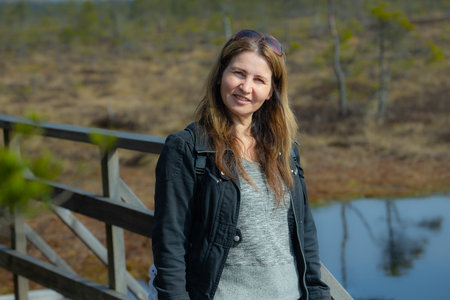 Portrait of a beautiful young woman with long brown hair, wearing a black jacket and sunglasses, standing on a wooden bridge in a swamp.の写真素材
