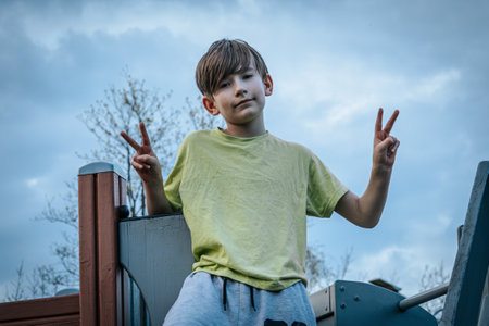 Little boy in a yellow T-shirt and blue jeans sitting on the playground and showing victory signの写真素材