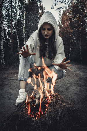 A beautiful girl in a white hood and jeans sits by the fire.の写真素材