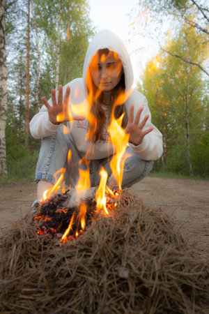 A girl in a hood sits at a bonfire in the forest.の写真素材