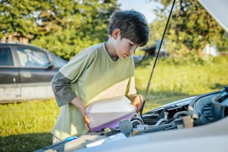young boy looking under hood of broken car with open hood on sunny dayの写真素材