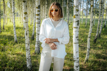 Portrait of a beautiful young woman in a white jacket and sunglasses on a background of birch trees.の写真素材
