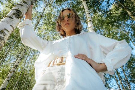 Portrait of a beautiful young woman in a white coat and sunglasses standing near a birch.の写真素材
