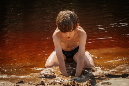 A boy in a black swimsuit sits on the shore of the river and looks at the water.